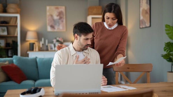 Shocked man looking at high internet or utility bill with partner offering support in a modern home